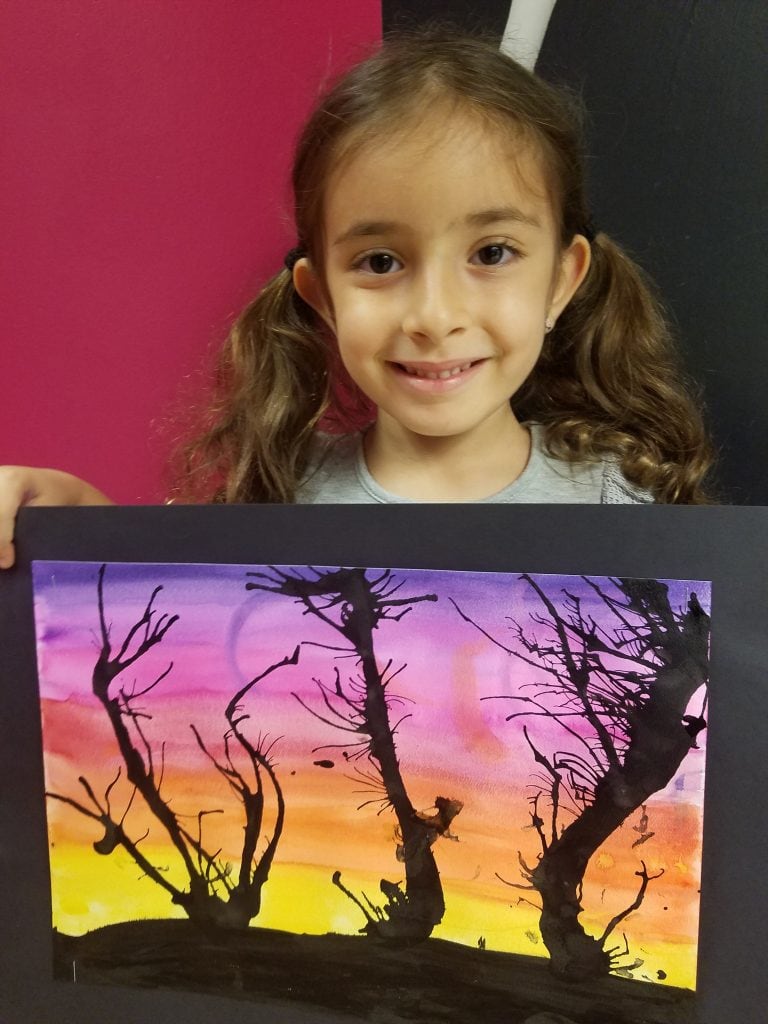 young girl with brown hair in pigtails holding water color sunset and silhouette trees