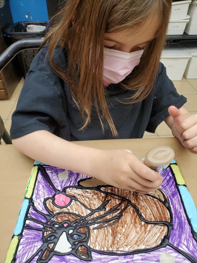 Young girl with brown hair wearing pink mask using black puffy paint to outline marker drawing