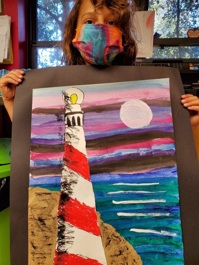 young girl wearing a tie dye masking holding water color painting of a lighthouse