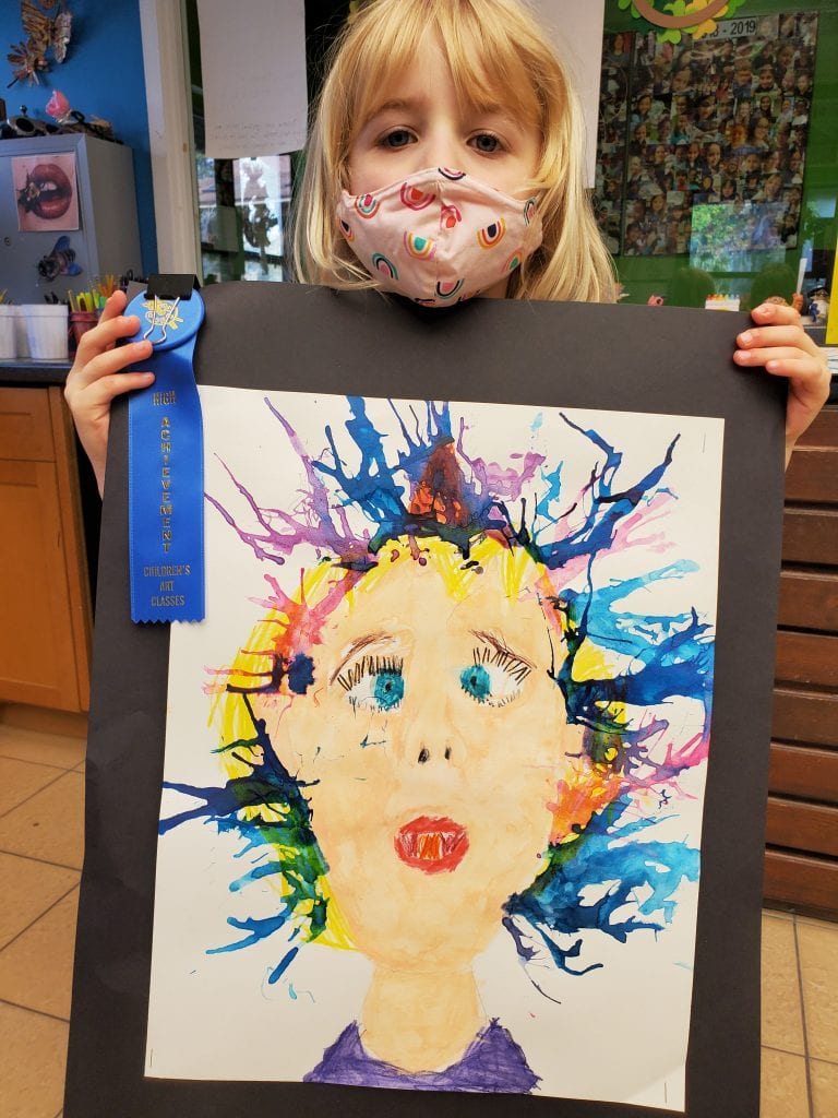 blonde young girl wearing pink mask with rainbows holding water color painting of women with colorful crazy hair and blue ribbon award
