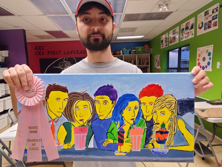 male student wearing hat holding acrylic painting of friends with a pink ribbon award