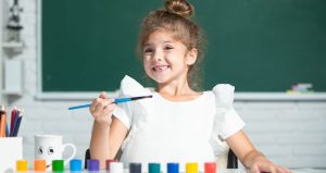 young girl in white shirt with hair pulled up in to a top bun smiling and holding a paint brush