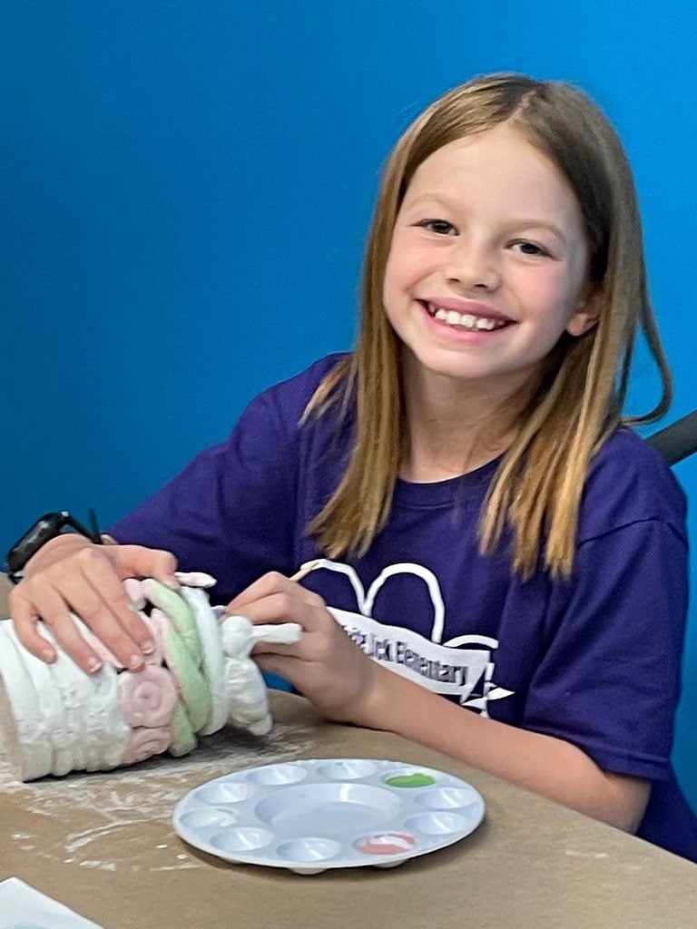 young girl in dark purple shirt sitting with a blue wall behind her making a ceramic coil pot