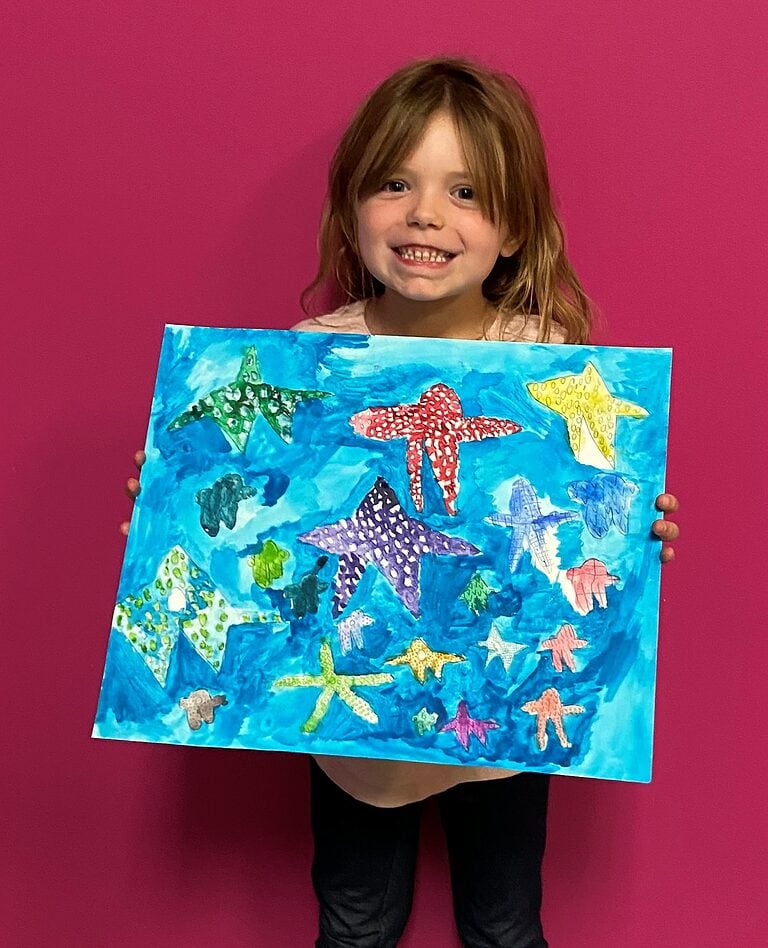little girl in black pants and peach top standing against a pink wall holding a starfish painting