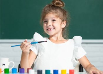 young girl in white shirt with hair pulled up in to a top bun smiling and holding a paint brush